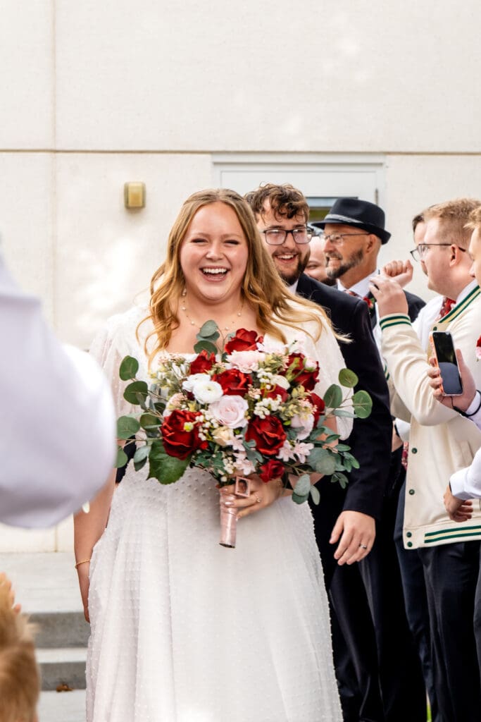 Bride smiles and holds bouquet as she walks through path of friends and family with groom behind her.