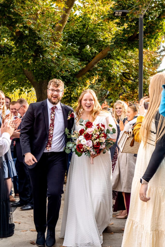 Bride and groom hold hands and smile big as they walk through path of friends and family who are cheering at the Idaho Falls Temple.