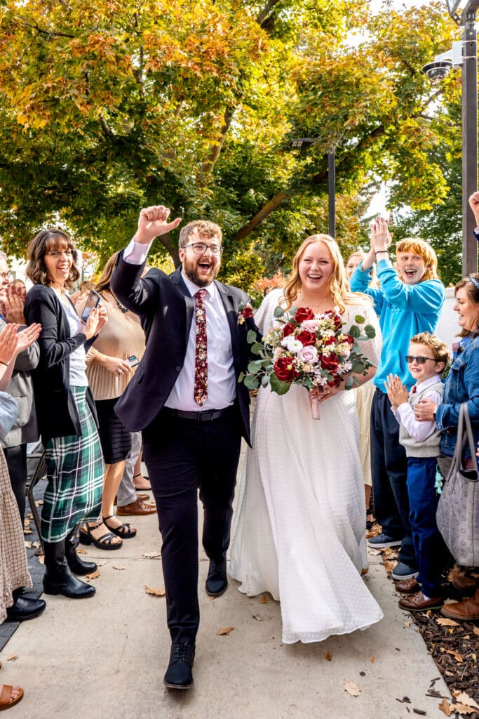 Bride and groom hold hands and walk through path of friends and family who are cheering as groom pumps fist in the air at the Idaho Falls Temple.