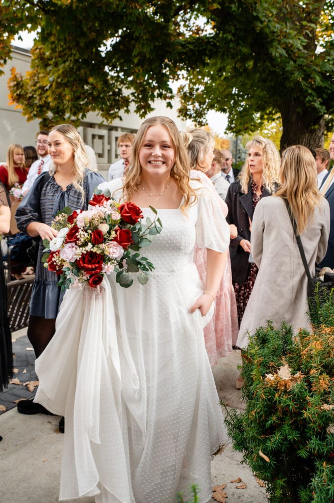 Bride smiles excitedly at camera while holding her train and bouquet in her right hands as family and friends mingle behind her at the Idaho Falls Temple.