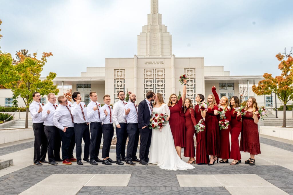Bridesmaids and groomsmen cheer as bride and groom kiss with beautiful blue skies and the Idaho Falls Temple behind them.