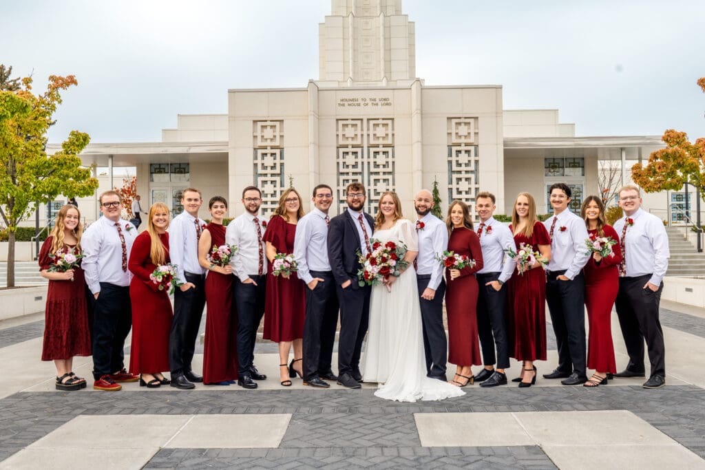 Bridesmaids and groomsmen alternate in a line and smile at camera with bride and groom with beautiful blue skies and the Idaho Falls Temple behind them.