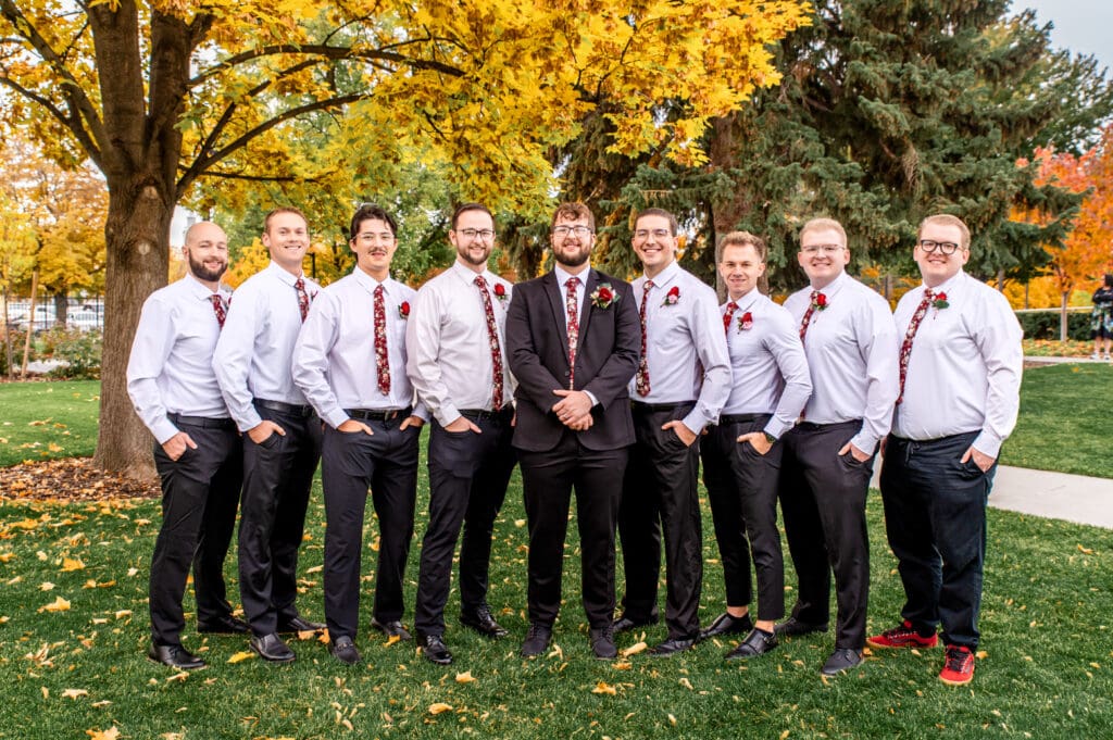 Groomsmen stand in line with hands in pocket and smile at camera with groom in front of fall colored trees at the Idaho Falls Temple.