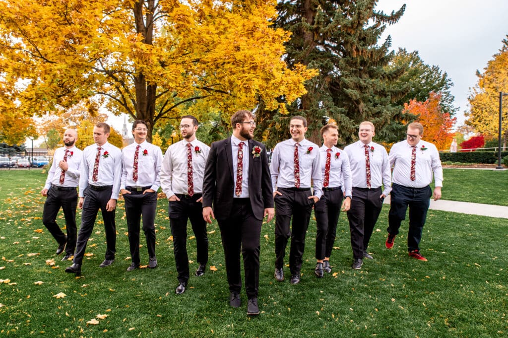Groomsmen walk in v line and smile with groom in front of fall colored trees at the Idaho Falls Temple.