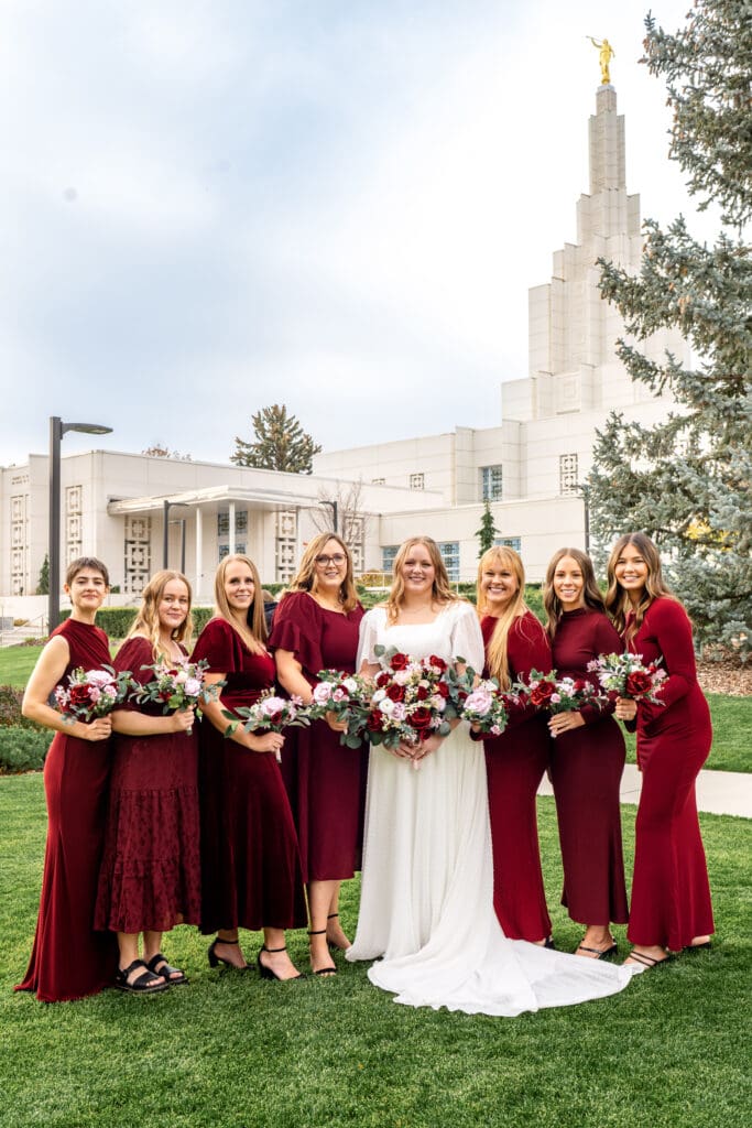Bridesmaids and bride stand in line, holding bouquets, and smile at camera in front of Idaho Falls temple.