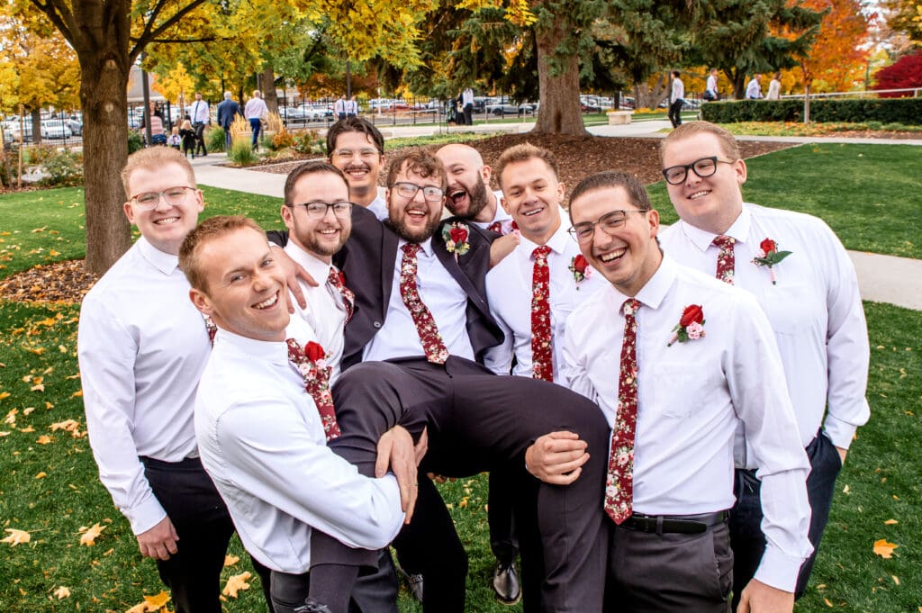 Groomsmen lift groom and smile at camera in front of fall colored trees at the Idaho Falls Temple.