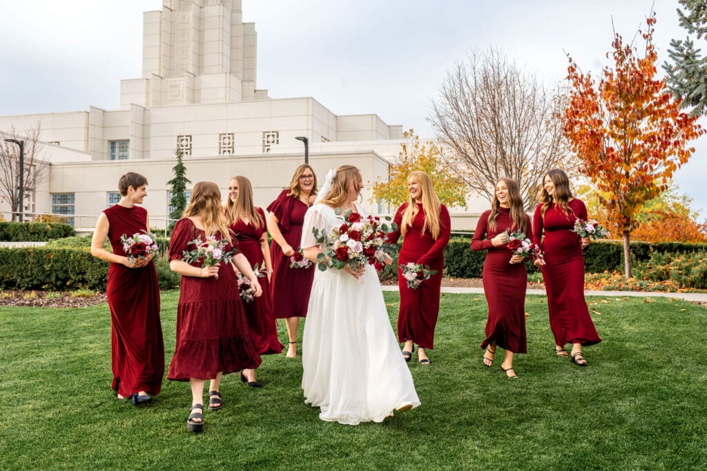 Bridesmaids and bride walk across grass and laugh with each other while holding bouquets in front of Idaho Falls temple.