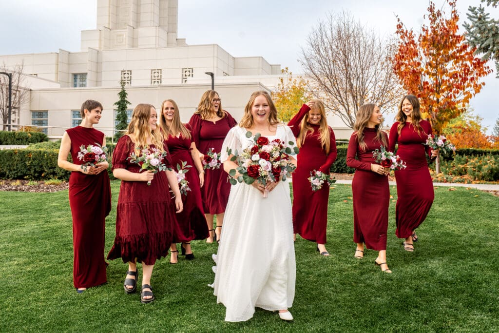 Bridesmaids and bride walk across grass and laugh while holding bouquets in front of Idaho Falls temple.