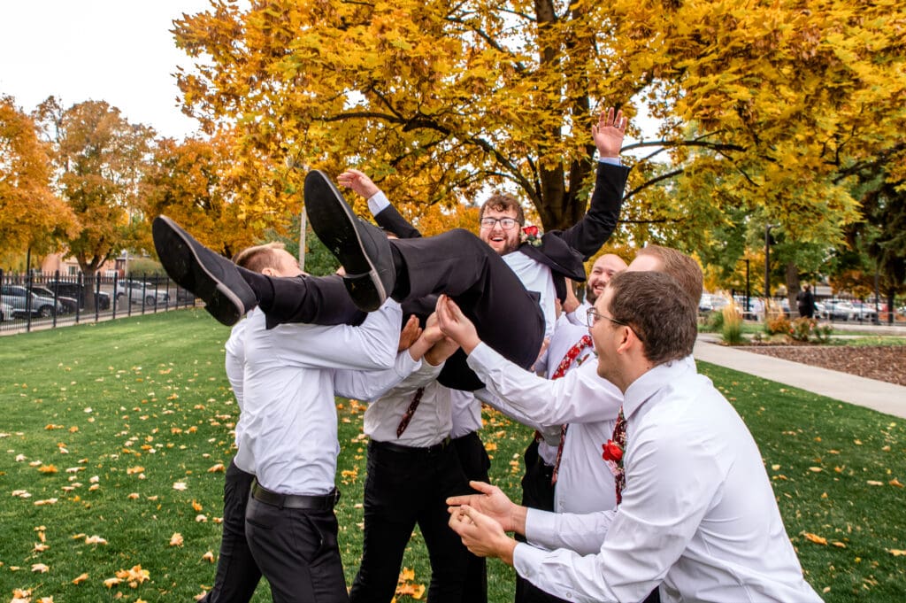 Groomsmen throw groom in air and laugh in front of fall colored trees at the Idaho Falls Temple.