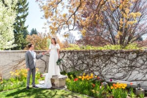 Bride stands on bench under blossoms with bouquet resting at her side as she takes groom's hand from below and smiles at him at Idaho Falls Temple.