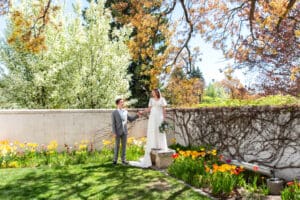 Bride stands on bench under blossoms with bouquet resting at her side as she takes groom's hand from below and smiles at him at Idaho Falls Temple, with beautiful spring flowers on the ground..