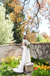 Bride under blossoms smiles down to her bouquet at her waist and stands on bench as bouquet rests at her side.