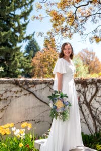 Bride under blossoms smiles over her should and stands on bench as bouquet rests at her side.
