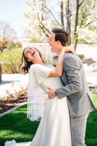 Bride leaning back laughing with eyes closed as her hands rest around groom's neck as groom leans in and laughs in front of blossoms at Idaho Falls Temple.