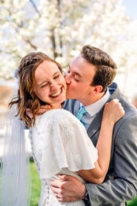 Groom bringing bride in by the waist and kissing her cheek as bride smiles with eyes closed and holds his suit collar in front of blossoms at Idaho Falls Temple.