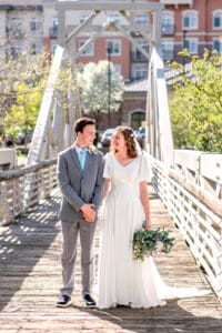 Bride and groom holding hands and smiling at each other while bride holds bouquet at her left side on bridge with city building background in Idaho Falls.