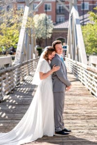 Bride leaning forward against groom's back and resting on his arm as groom stands straight and the both close their eyes on bridge with city building background in Idaho Falls.