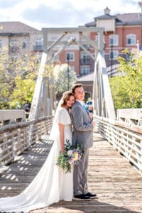 Bride leaning forward against groom's back, resting the bouquet by her side as groom stand straight and holds his suit and they both smile at the camera on bridge with city building background in Idaho Falls.