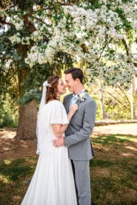 Bride and groom standing under blossoms and smiling at each other with noses touching, with groom holding bride at the hips and bride resting hands on groom's chest in Idaho Falls.