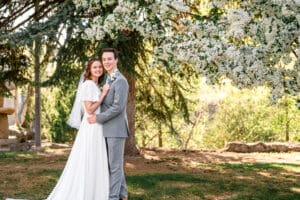 Bride and groom standing under blossoms and smiling at the camera with groom holding bride at the hips and bride resting hands on groom's chest in Idaho Falls.
