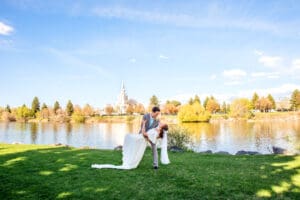 Bride and groom smiling at each other as groom dips bride on grass in front Snake River and the Idaho Falls Temple.