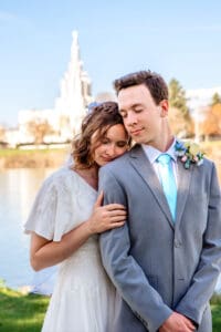 Bride standing behind groom and resting her head on his shoulder and her hand on his arm in front of snake river and Idaho Falls Temple.