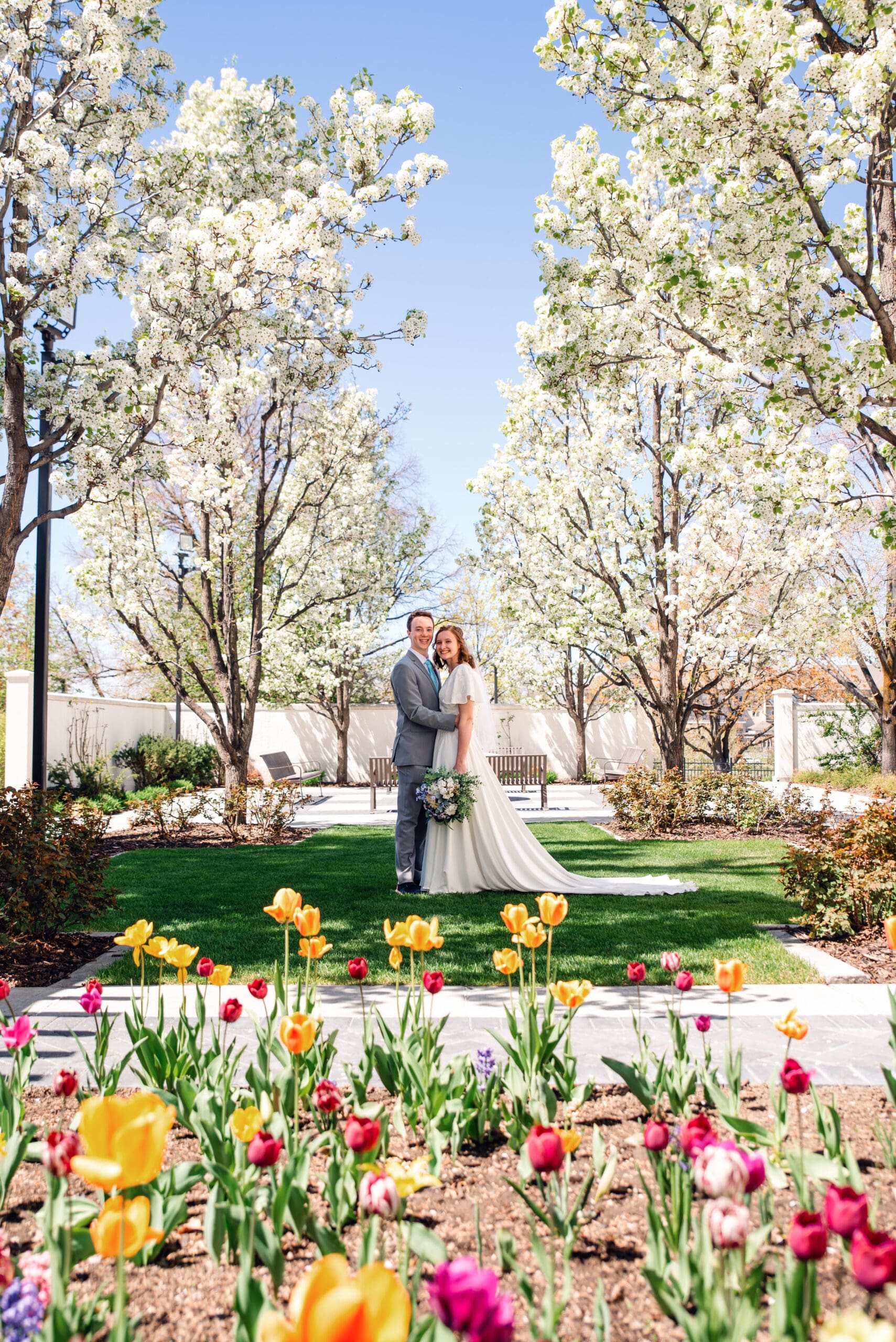 Bride and groom facing and smiling at camera as bride holds bouquet at her side with the couple framed by blossom trees and colorful flowers at the Idaho Falls Temple.