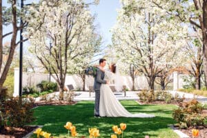 Bride and groom facing each other and with bride wrapping her arms around groom's neck with bouquet with the couple framed by blossom trees and colorful flowers at the Idaho Falls Temple.