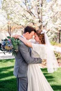 Bride and groom facing each other and touching foreheads with bride wrapping her arms around groom's neck with bouquet in front of blossom trees at the Idaho Falls Temple.