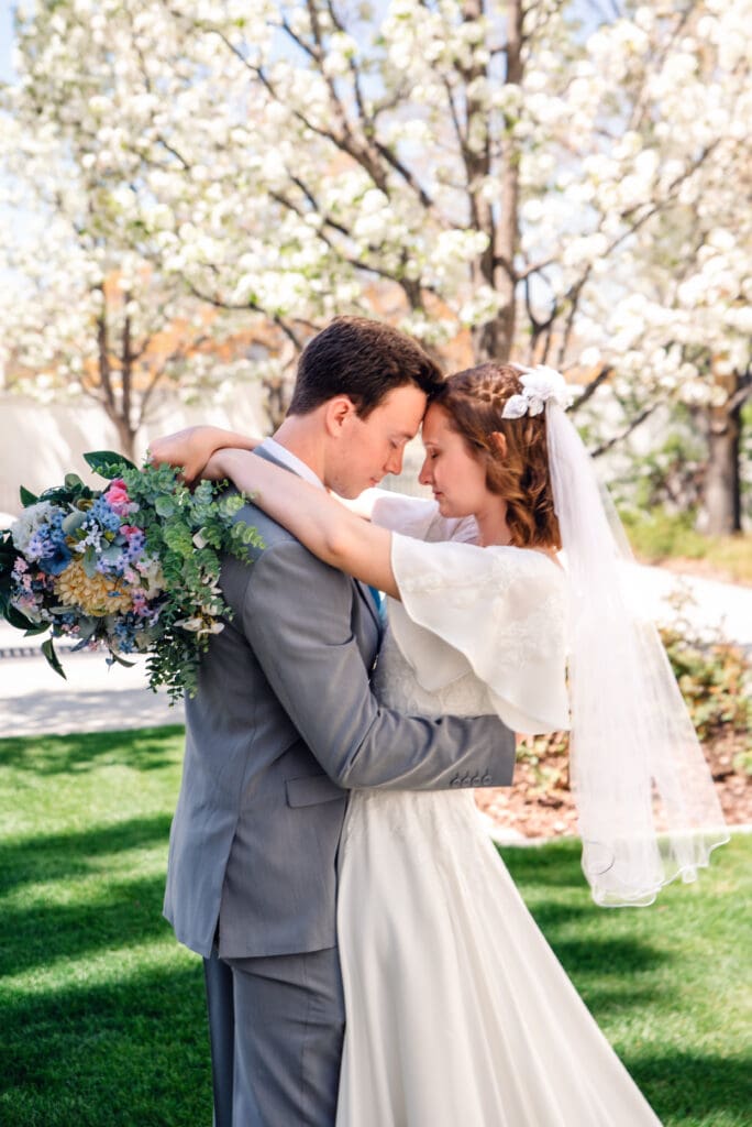 Bride and groom facing each other and touching foreheads with bride wrapping her arms around groom's neck with bouquet in front of blossom trees at the Idaho Falls Temple.