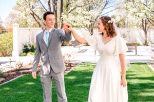 Bride and groom smile at each other as groom leads bride by hand in dance and bride holds train in front of blossoms at the Idaho Falls Temple.