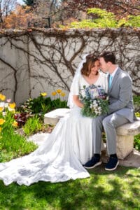 Bride and groom sitting on bench under blossoms as they smile and touch groom's nose touches bride's cheek with closed eyes as bouquet sits on their laps and dress train drapes down at Idaho Falls temple.