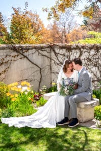 Bride and groom sitting on bench under blossoms as they smile and touch foreheads with closed eyes as bouquet sits on their laps and dress train drapes down at Idaho Falls temple.