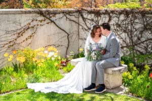 Bride and groom sitting on bench under blossoms as bride smiles and they touch foreheads with closed eyes as bouquet sits on their laps and dress train drapes down at Idaho Falls temple.