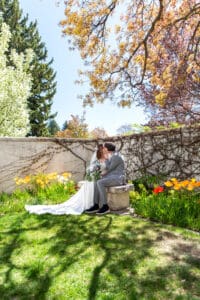Bride and groom sitting on bench under blossoms as groom brings bride in for kiss and bouquet rests on their laps at Idaho Falls Temple.