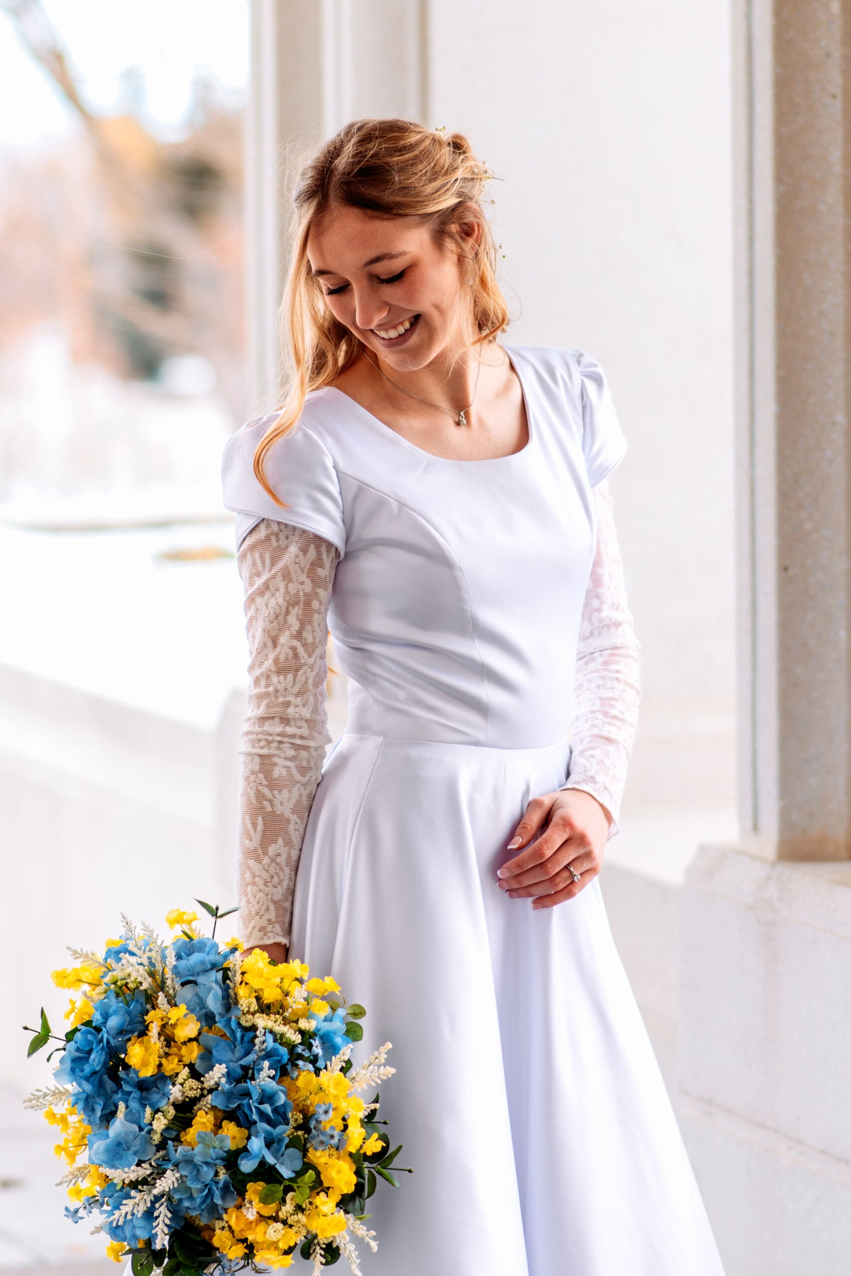 Bride in wedding dress, smiling and looking down her arm at her yellow and blue bouquet at the Rexburg Temple.