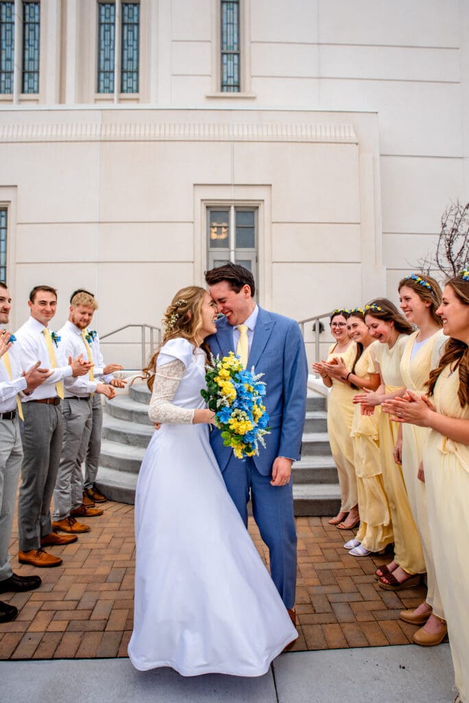Bride and groom hugging in front of temple exit steps with bouquet and wedding party surrounding them at the Rexburg Temple.