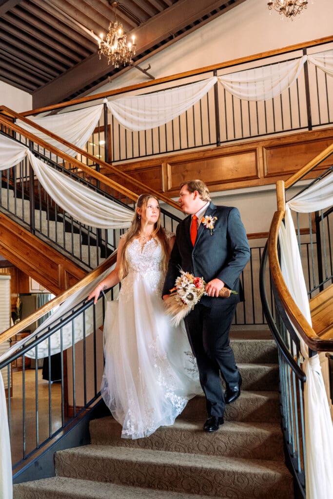 Bride and groom walk down carpeted stairs with decorated wooded banisters while holding hands and smiling at each other at The Atrium, captured by Rexburg wedding photographer.