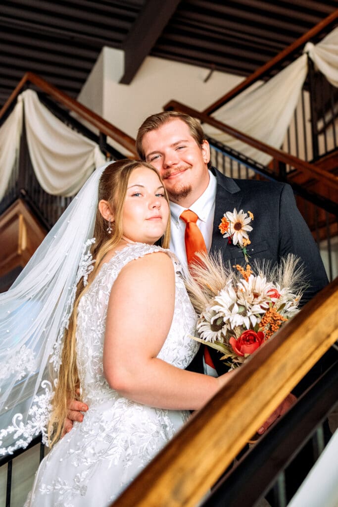 Bride and groom smile at camera as they stand on stairs with bride holding bouquet at venue in Rexburg, Idaho. 