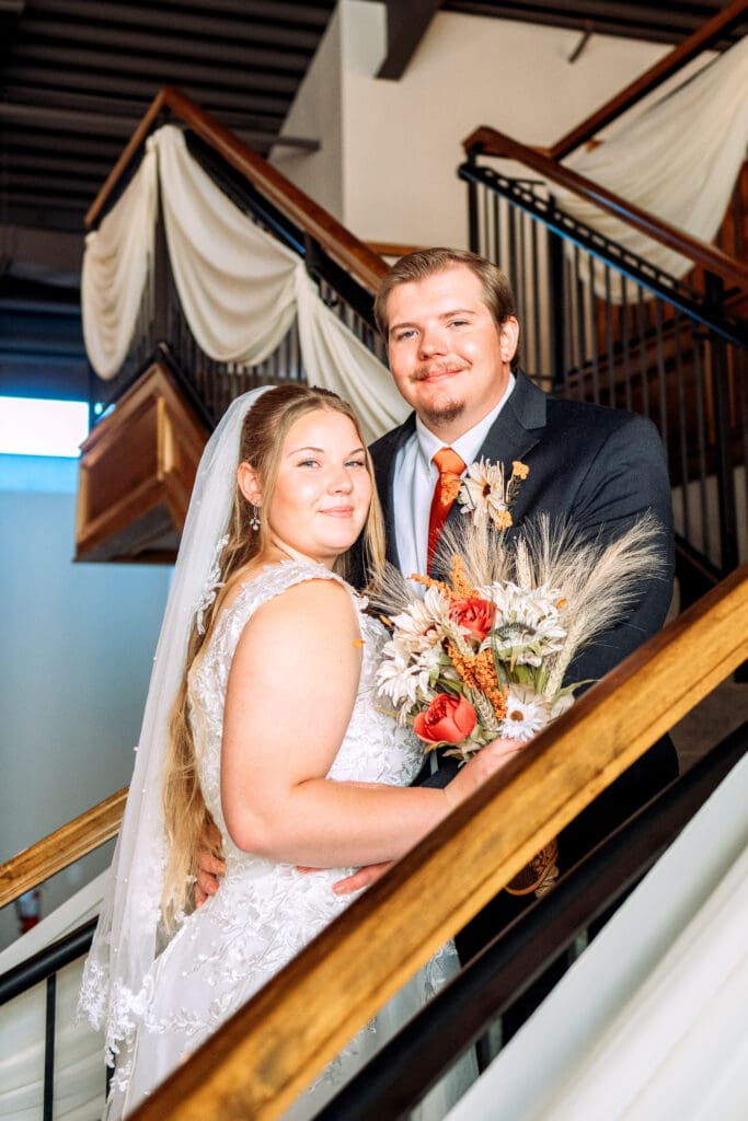 Bride and groom face each other on stairs and smile at camera while bride holds bouquet with wooded banisters in the front and backgrounds at The Atrium, captured by Rexburg wedding photographer.