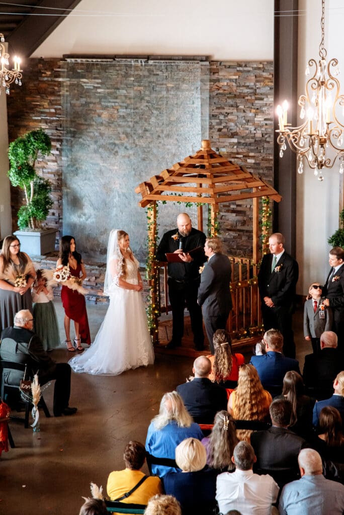 Bride and groom smile at each other in front of officiant while family and friends watch with waterfall and gazebo in back at The Atrium, captured by Rexburg wedding photographer.
