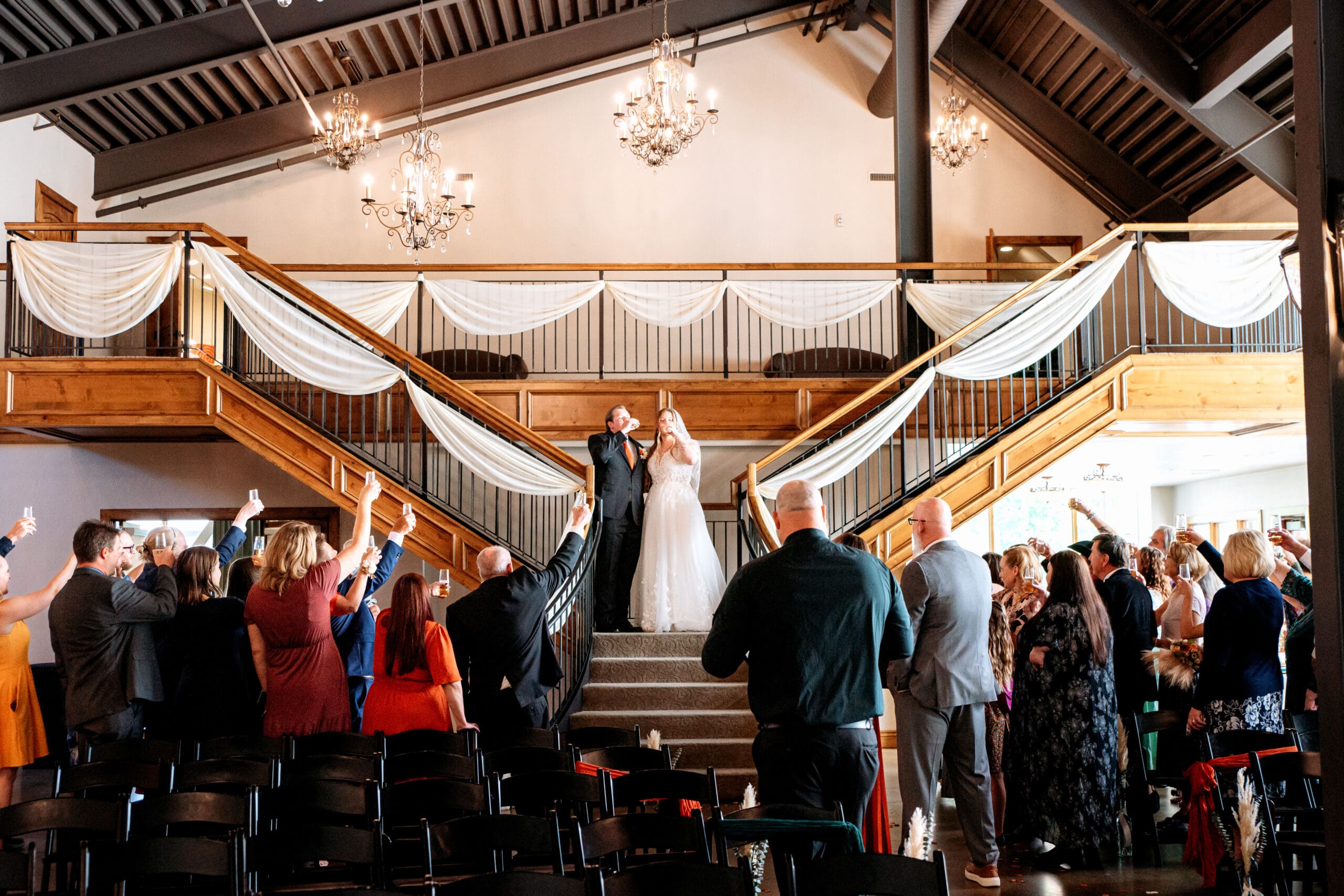 Bride and groom sip champagne while standing on top of decorated stairs as friends and family raise glasses to toast them at The Atrium, captured by Rexburg wedding photographer.