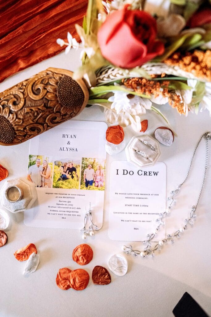 Bouquet in custom wood holder, red and orange flower petals, wedding invitations and announcements, bride's rings and jewelry and orange table runner, captured by Rexburg wedding photographer.