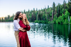 Man wraps arms around woman's waist they smile and stare off into the distance as they stand in lake with trees in back, captured by Utah engagement photographer.