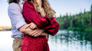 Man wraps arms around woman's waist and woman in red dress wraps her arms around his, with the both of them showing off their jewelry, captured by Utah engagement photographer.