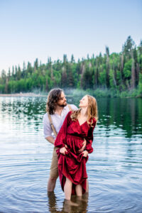 Man and woman in red dress stand in lake as man holds woman's hips and woman holds her dress and the two smile at each other, captured by Utah engagement photographer.