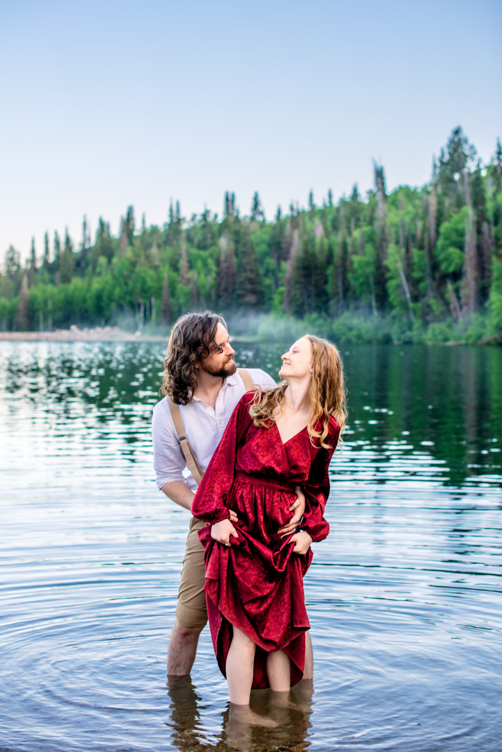 Man and woman in red dress stand in lake as man holds woman's hips and woman holds her dress and the two smile at each other, captured by Utah engagement photographer.