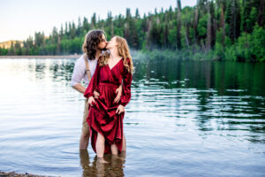 Man places hands on woman's hips while woman holds skirts of dress and the two kiss and stand in lake with trees in back, captured by engagement photographer.