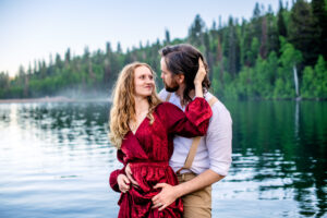Man holds skirts of woman's dress as woman places hand on his head and smiles at him with them standing in a lake, captured by Utah engagement photographer.