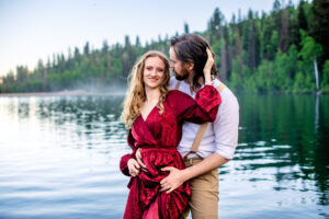 Man holds skirts of woman's dress as woman places hand on his head and smiles at camera with them standing in a lake, captured by Utah engagement photographer.